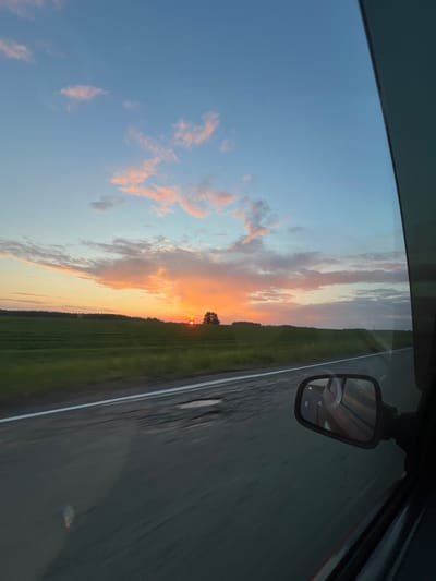 The image captures a sunset view from inside a vehicle traveling on a road. The sky is painted with hues of orange, pink, and blue, with scattered clouds. A distant treeline borders a green field. In the foreground, the road surface is visible, showing some wear and tear, with a white line marking the lane. A side mirror reflects the car's interior and the sunset. The scene is set in Sidorovy Gory, Russia, suggesting a rural or suburban setting during twilight. There are no people or specific actions depicted, focusing solely on the landscape and the atmospheric conditions of the sunset.