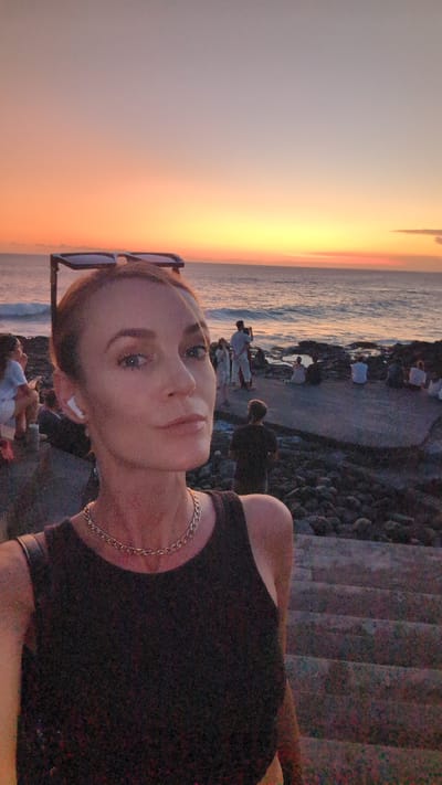 The image shows a woman posing for a selfie in Pererenan, Indonesia, at sunset. She has fair skin, blue eyes, and her hair is tied back. She's wearing a black sleeveless top and a silver chain necklace, with sunglasses perched on her head. To her right, the ocean reflects the vibrant orange and yellow hues of the setting sun. In the background, there's a walkway with people enjoying the sunset, and to her left, there are steps leading down to the area. The scene captures a moment of leisure and the beauty of the natural surroundings.