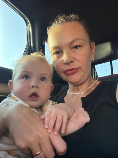 In Koliushevo, Russia, a woman and a baby are pictured in what appears to be the interior of a vehicle. The woman is in the foreground, holding the baby. The baby, with blonde hair and wide blue eyes, is looking upwards, with its mouth slightly open. The woman has her hair pulled back and is wearing a black top and a white beaded necklace with a heart pendant. The environment suggests they are traveling, possibly during daylight hours, given the light coming from the car window.