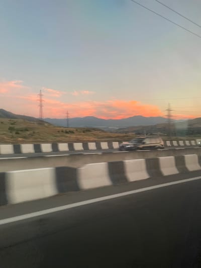 The image captures a sunset scene on a highway likely near Dzveli Kanda, Georgia, as indicated by the provided context. The sky is painted with hues of pink and orange, with scattered clouds. In the distance, rolling hills and mountains form the backdrop. Two tall, lattice-like power transmission towers are visible on the left side of the frame. A highway with a clear white line marking the lane runs through the foreground. A concrete barrier, alternating between white and dark gray sections, separates the highway lanes. A dark-colored SUV is seen driving on the highway, its headlights on, indicating it's either dusk or early morning. The overall impression is that of a landscape viewed from a moving vehicle on a highway.