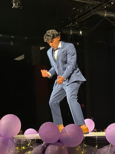 A male individual stands centered on a decorated theatrical stage in Palos Verdes Estates, United States, captured in a dynamic, mid-action pose. He is wearing a light blue three-piece suit, a white dress shirt, a dark bow tie, and tan dress shoes. His dark, wavy hair is styled upwards, and he wears a wristwatch on his left wrist. Both hands are clenched into fists and held near his chest, while his mouth is open. The immediate foreground of the stage is adorned with numerous matte purple balloons, purple mesh fabric, and small string lights. The background is dark, revealing overhead stage lighting fixtures, cables, and numbered structural beams (e.g., '79', '78'). Faint outlines of spectators are visible in the lower right background.