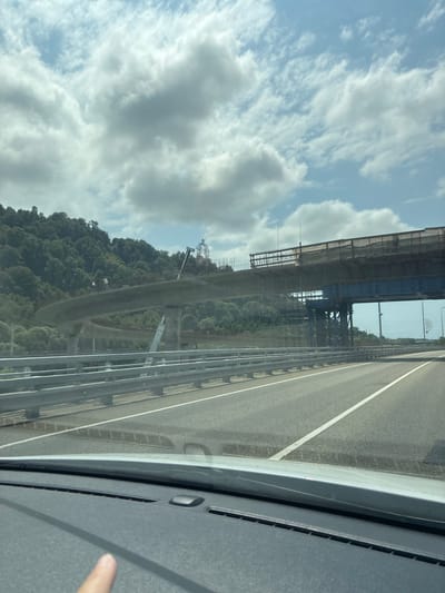 The image depicts a highway under construction, with a large, partially built elevated road structure dominating the scene. The road appears to curve into the distance, supported by concrete pillars and scaffolding. Construction equipment, including cranes and workers, are visible on and around the structure. To the left of the construction, a lush, tree-covered hillside rises steeply. The highway itself has multiple lanes marked with white lines, and a metal guardrail runs along the side. The sky above is partly cloudy, with scattered white clouds against a blue backdrop. The foreground shows the dashboard of a vehicle, with a finger pointing towards the scene outside. The image does not contain any information about a specific city like Vysokoye, Russia.
