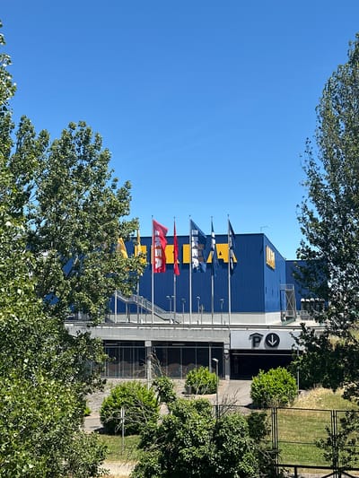 The image depicts the IKEA store located in the city of Oviedo, Spain. The facade of the store is predominantly blue, with large yellow IKEA logos visible on the upper portion. A series of flags, including red and yellow ones with the IKEA logo, are flying in front of the building. Stairs lead up to an entrance. Below the main store, there is a section with windows and an entrance to a parking garage, indicated by a "P" symbol with a downward arrow. The scene is framed by lush green trees on the left and right sides, and the sky above is clear and blue. The lighting suggests a bright, sunny day.