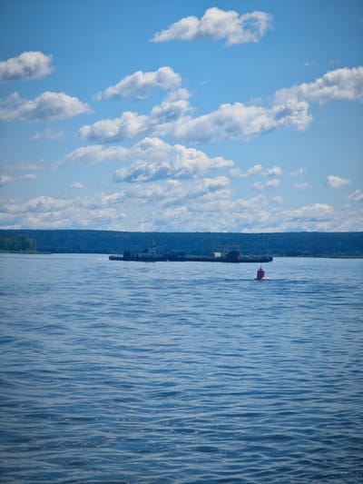 The image captures a wide view of a calm body of water, likely a river, on a clear day. The sky above is a vibrant blue, dotted with fluffy white cumulus clouds. In the distance, a dense, dark green forest lines the horizon.
On the water, a long, dark-colored barge is visible, moving from left to right. A small red buoy floats in the foreground, closer to the viewer than the barge. The water itself is a deep blue, with gentle ripples creating a textured surface. The overall impression is one of peacefulness and natural beauty. There are no people visible in the image. The location is Krutoi Log, Russia.
