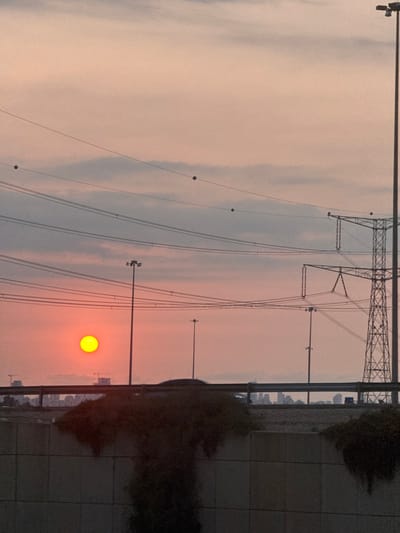 The image depicts an urban-suburban landscape at either sunrise or sunset, originating from the city of Rosh Haayin, Israel. A distinct, bright yellow-orange sun is positioned low on the left-center horizon. The sky exhibits varying shades of pale orange, pink, and light grey, indicative of dawn or dusk, with scattered cloud formations. Multiple dark-colored power lines are visible, stretching horizontally across the upper and middle portions of the frame; some lines feature small, dark spherical markers, likely for aerial visibility. Several tall, dark utility poles, equipped with multiple luminaires, punctuate the middle ground. A large, dark lattice-steel electricity transmission tower stands prominently on the far right side of the frame. In the immediate foreground, a concrete barrier or retaining wall is visible, partially covered by sparse, dark vegetation. Above this barrier, an elevated road or highway is discernible, marked by a metallic guardrail. Faint silhouettes of vehicles can be observed on the road, suggesting ongoing traffic. In the distant background, a cityscape with numerous buildings and visible construction cranes forms a silhouette against the horizon. No specific individuals or explicit interactions are identifiable within the scene.