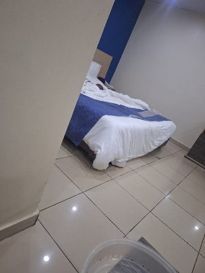 This image depicts an interior room, located in Close, Nigeria. A single individual is present, lying supine in a bed with a light-toned wooden headboard. The person is largely obscured by white bedding, with only the upper portion of their head visible against a white pillow. The bed features a blue bedspread beneath the white duvet. An unidentified small object rests on a surface immediately adjacent to the pillow. The room's walls are painted cream in the foreground, transitioning to a two-toned blue and white scheme behind the bed. The floor consists of glossy, light-colored square tiles with dark grout lines, reflecting ambient light. A translucent plastic bucket, partially visible in the bottom right, contains several dark, folded textile items.