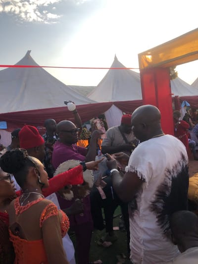 An outdoor daytime gathering is depicted in Amanuke, Nigeria, featuring multiple individuals engaged in handling Nigerian currency notes. In the immediate foreground, a dark-skinned man with a bald head, wearing a white short-sleeved top with a black tie-dye pattern and frayed edges, is observed from a rear-right angle, meticulously organizing or counting currency notes in his hands. A gold chain is visible around his neck and a silver watch on his left wrist. To his left, a dark-skinned man in glasses and a purple short-sleeved shirt holds a bundle of Nigerian Naira notes extended forward in his right hand, while his left hand holds a white spherical object raised above his head. Further left, a woman with an intricate updo, wearing an orange and sheer top adorned with beads and sequins, is seen from the side-back, pointing her right arm forward. She holds a white and brown traditional hand fan in her left hand. Scattered Nigerian Naira notes are visible on the ground beneath the central figures. In the background, several other attendees are visible, some wearing traditional red caps. The scene is set against a backdrop of multiple white conical tents adorned with red fabric drapes and a horizontal red ribbon or line spanning the upper mid-ground. A decorative red and yellow archway is partially visible on the far right. The sky overhead is bright with visible cloud formations.