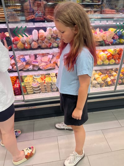 This is a vertical, eye-level photograph taken inside a brightly lit grocery store in Navahrudak, Belarus. The focus is on a young girl with long, blonde hair streaked with pink, looking down at a refrigerated display case filled with various processed meats and dairy products. She is wearing a light blue t-shirt and a dark skirt. Her left arm is visible, and she appears to be holding a phone with a red case in her right hand, which is partially obscured.

In the foreground, the legs and feet of another person are visible. This person is wearing a white top and dark shorts, and decorated slides on their feet. The other person's feet are wearing slippers decorated with cartoon characters, and one foot is reaching out towards the grocery display. The floor of the store is tiled with light grey squares.

The refrigerated display case is packed with a variety of packaged food items, including sausages, hams, cheeses, and other dairy products. Some of the visible text on the packaging and labels includes "XX", "4 р 43 к", "7067 6028", "Гр 28 к 5 р 27", "Залив", "МАЛАДЗЕЧНА", "Б", and "МОЛОЧНЫЕ". The overall impression is a casual shopping trip, with the child focused on the food items in the display. The lighting suggests it is daytime, and the environment is clean and well-stocked.