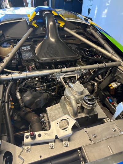 The image shows the engine bay of a race car, specifically a Lamborghini Huracan Super Trofeo EVO2. The view is from underneath the car, looking upwards. The engine is visible, surrounded by the car's chassis and various components such as the exhaust system, cooling system, and wiring harnesses. The location is likely a pit garage at the Vallelunga Circuit near Campagnano di Roma, Italy, a common venue for racing events and testing of race cars like the Super Trofeo.