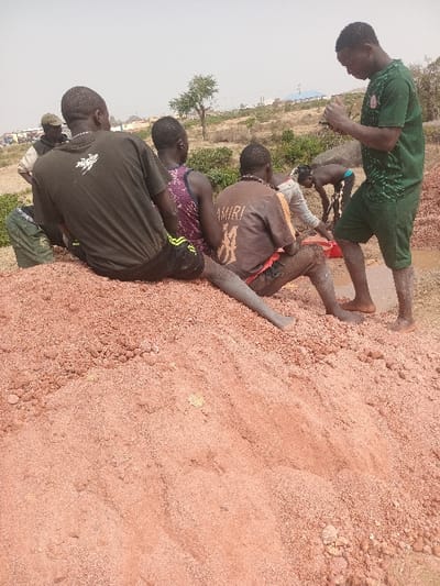 A group of young men are situated on a reddish-brown, sandy terrain. They are dressed in casual wear, including shorts and t-shirts of various colors like green, black, and brown. The landscape is relatively arid, with some greenery visible in the background, including a few trees and shrubs. A hazy sky hangs overhead. The overall scene evokes a sense of casual interaction and outdoor activity within a natural setting.