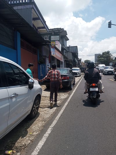 The photograph captures a street scene with parked vehicles and pedestrian activity. A white car is parked on the left side of the frame, with two men standing near it. One man, wearing a green shirt, is closer to the car, while the other, dressed in a red and black plaid shirt and brown pants, is walking away from the vehicle. To the right, a black Honda City is parked behind the white car. A scooter with two riders is traveling down the road. Traffic is visible in the background, with multiple cars lined up on the road. Buildings line the street, and a sign reading "TOKO AR ROHMAH" is displayed on one of them. The sky is partly cloudy. The location of the photograph is in a city. The name "Kioumaknao 2" is not visible in the photograph.