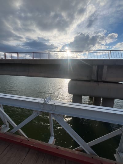 This image, apparently taken in Ait Mazigh, Morocco, shows a view looking down from a bridge. The picture is oriented with the sky on the left and water on the right. The sky is filled with cumulus clouds and the sun is visible. A bridge structure is visible in the center. A white metal railing runs along the edge of the bridge. The water below is green. Steel support beams of the bridge are present. Wooden boards are seen at the bottom right.
