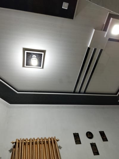 The image shows a close-up view of a modern interior ceiling with recessed lighting and decorative elements. The ceiling is multi-layered and features a combination of white and black panels. One recessed light fixture is square with a black border and a speckled interior, emitting a bright white light. Another light fixture on the right is also square and illuminated. Decorative black stripes are incorporated into the ceiling design, creating a geometric pattern. The border of the ceiling features a black textured band with white trim. Below the ceiling, a wall is visible, displaying several framed Islamic calligraphy art pieces and a circular clock with Arabic script. On the left side of the wall, a set of gold-colored curtains with ornate tie-backs is partially visible. The overall impression is of a contemporary and well-decorated indoor space.