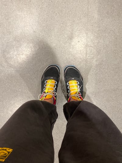 The media file shows a top-down view of a person's feet wearing black athletic shoes with bright yellow accents and white laces. The shoes have a mesh upper and appear to be well-worn. The person is wearing dark-colored sweatpants. The ground is a light grey, speckled linoleum floor with visible seams and some scuff marks, suggesting an indoor environment.
The image is taken from a high angle, looking down at the feet. There is no discernible activity or event taking place; the scene appears to be a static shot. The lighting suggests an indoor setting, possibly a gym, school, or public building, with overhead lighting. There are no obvious weather cues or time-of-day indicators.
No specific emotions are conveyed. The setting is generic and does not provide strong location cues beyond being indoors. There is no visible text in the image, except for a small, unreadable logo on the side of the shoe.
The overall impression is a simple, everyday scene focusing on footwear and casual attire. The location context of Uppsala, Sweden, does not contribute any specific details observable within the image itself.