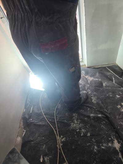 A first-person perspective image shows a person standing in what appears to be an interior room undergoing renovation, possibly in Gorno Aleksandrovo, Bulgaria. The person is wearing dark work pants with a red patch on the thigh and grey shoes. The floor is covered with black plastic sheeting, which is splattered with white material, suggesting plaster or paint. The room's walls are painted in light grey. A white electrical cord runs along the floor near the right side of the image. The light source is bright, obscuring some of the details.