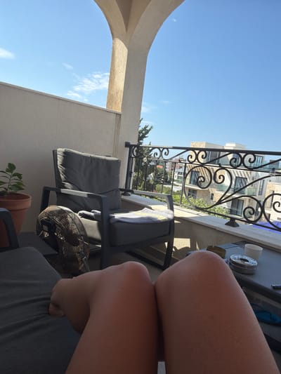 The image shows a first-person perspective on a balcony in Dobra Voda, Montenegro. The foreground is dominated by a person's legs, suggesting they are sitting or reclining. In the mid-ground, a grey armchair is positioned on the balcony, with a fan placed in front of it. A small potted plant is visible to the left of the armchair. Beyond the balcony, an ornate black metal railing overlooks a cityscape with buildings and trees under a clear blue sky. The scene appears to be set during daytime, with sunlight casting shadows.