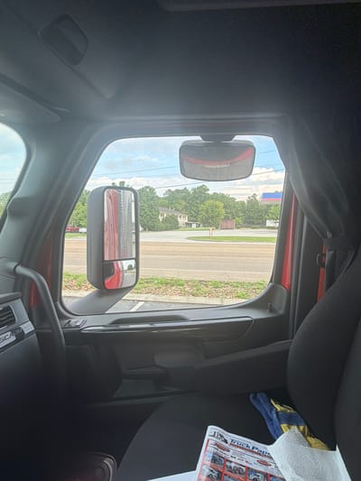 The image is taken from inside a truck cab, looking out the driver's side window. The truck appears to be stopped, possibly at an intersection or parking lot. Outside the window, a road is visible, with a sidewalk and a grassy area. Beyond that, there are trees, buildings, and a gas station with a red and blue sign. In the foreground, inside the truck, a newspaper titled "Truck Paper" is visible on the passenger seat, along with a pair of work gloves. The sky is cloudy. No people are visible in the image. The location is identified as Chattanooga, United States of America.