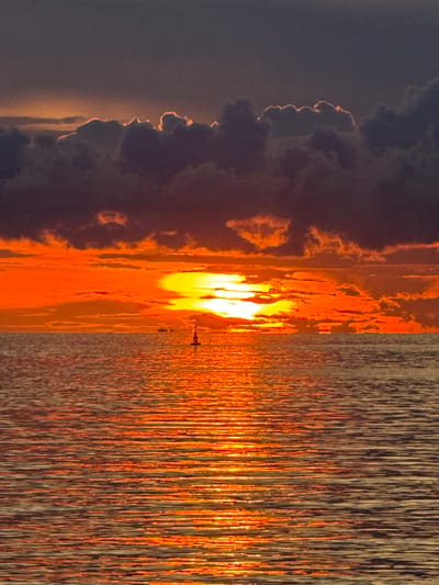 The image captures a sunset over water, likely the sea or a large body of water, in Baan Wok Tum, Thailand. The sun, a brilliant, intense white, is positioned centrally in the frame, with a vertical orange reflection radiating across the water's surface. Dark, textured clouds occupy the left side of the image, the edges of which are illuminated by a soft, orange glow from the setting sun. The water displays an intricate pattern of vertical lines, indicating wave movement and light reflection. There is an indication of an object in the water, possibly a boat.