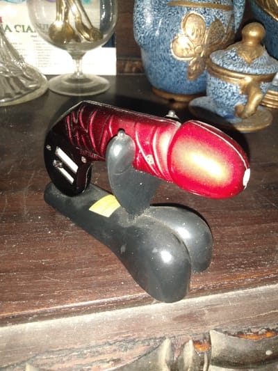The image displays a close-up of a variety of objects arranged on a dark, polished wooden surface, consistent with a shelf or tabletop. The surface shows visible dust accumulation, particularly along its carved front edge.
In the foreground, a novelty item is positioned horizontally. This object features a glossy red, rounded tip, a metallic red body with a textured, possibly ribbed, pattern towards its base, and a black, bifurcated base that supports the item on the surface. A small yellow label is affixed to the black base. Silver-colored details, possibly screws or decorative studs, are visible on both the black and red sections of the item.
In the background, from left to right, are a partially visible clear glass decanter, a clear stemmed glass containing several small, golden-colored spoons, and a blue speckled ceramic pot with a golden, butterfly-like embellishment. To the right of this pot is a smaller, matching blue speckled ceramic lidded container with golden accents, resembling a miniature teacup. A piece of paper or card, with text partially visible (e.g., "A CIA"), stands upright behind the stemmed glass.
No individuals, actions, or interactions are depicted. The lighting suggests an indoor setting. The location in the city of Косшы, Kazakhstan, cannot be visually confirmed from the details within the image.