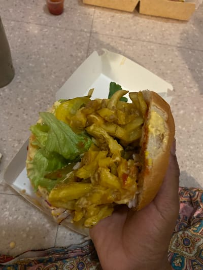 A hand holds a sandwich over a white cardboard container. The sandwich is composed of a bun, lettuce, fried potatoes, and sauces. The background consists of flooring and a patterned fabric. The location is not specified as Le Pré-Saint-Gervais, France in the image.
