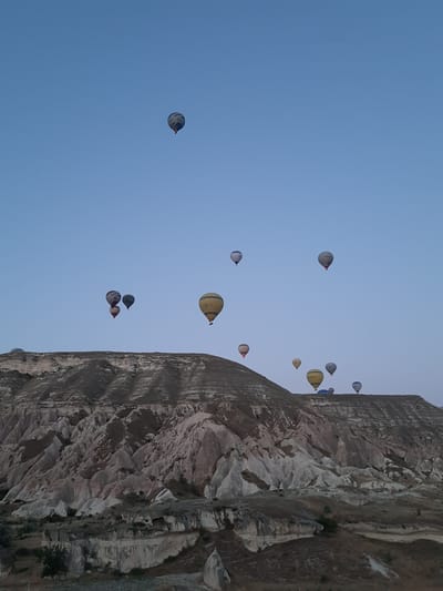 This image depicts a scenic view of hot air balloons ascending over a rocky, eroded landscape, likely in or near Avanos, Turkey, a region renowned for its hot air ballooning activities. The sky is a clear, pale blue, indicating either early morning or late evening. Multiple hot air balloons of various colors and sizes are scattered across the sky, some appearing close to the ground and others higher up. The terrain below consists of rugged, undulating hills and rock formations with visible textures and striations, indicative of geological weathering. The foreground shows dry, sparse vegetation and some smaller rock outcrops. The overall atmosphere is tranquil and vast, highlighting the interplay between the natural landscape and the aerial spectacle of the balloons.