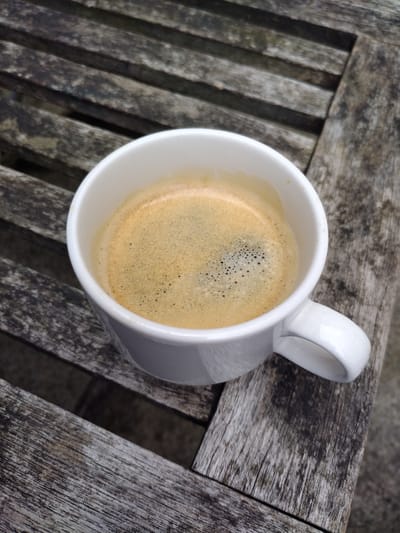 A white ceramic mug filled with freshly brewed coffee sits on a weathered wooden table. The coffee has a rich crema, with small bubbles dotting the surface, indicating it might be an espresso or a similar coffee drink. The mug has a simple, classic design with a curved handle.
The table's surface is made of wooden planks, showing signs of age and exposure to the elements, with a textured, slightly worn appearance. The planks are oriented horizontally in the image. The lighting suggests it is daytime, likely outdoors, given the natural texture and potential outdoor setting of the table. The overall mood is one of a quiet, perhaps solitary moment of enjoying a hot beverage. There are no people visible in the image. The setting appears to be a relaxed, casual environment.