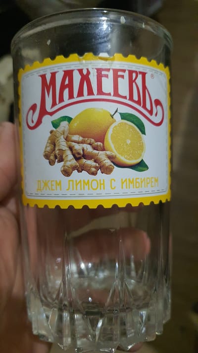 A close-up, eye-level shot shows a glass jar with a label that reads "МАХЕЕВЪ" in red lettering. Below the brand name are illustrations of lemons and ginger roots, with the text "ДЖЕМ ЛИМОН С ИМБИРЕМ" in yellow lettering. The jar is partially filled with a clear liquid. The background is blurred and appears to be a dimly lit indoor setting. The image does not provide any information that would suggest a location in Karankul, Uzbekistan, or any other specific city.
