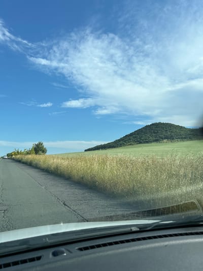 This image depicts a scenic landscape, likely from the perspective of someone in a vehicle traveling along a rural road. The foreground shows the cracked asphalt of the road and the dashboard of a car. To the right of the road, tall, dry grass lines the verge, leading to a vast, green field. In the distance, a large, conical hill covered in lush green trees dominates the horizon. The sky is a vibrant blue, with scattered white clouds. A few trees are visible on the left side of the road, near the edge of the grassy field. The overall impression is one of a bright, sunny day in a rural setting. The image does not provide specific details that would place it in the city of Medovo, Bulgaria.