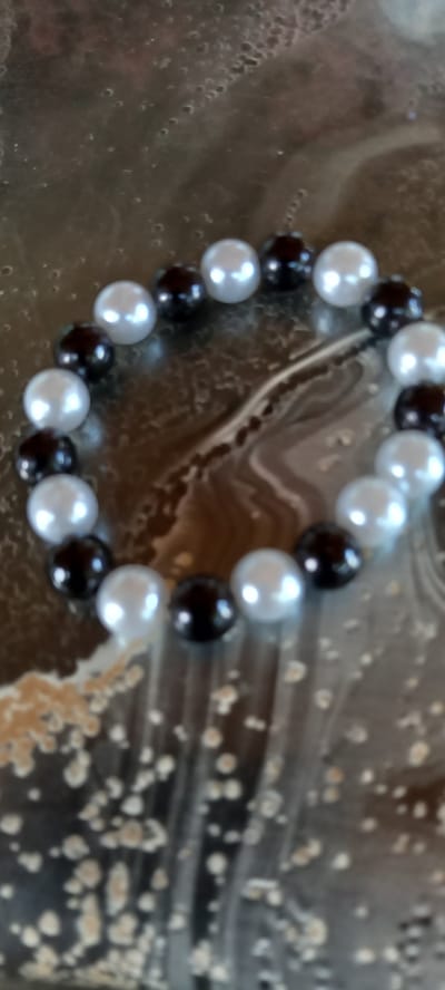 The image is a close-up, overhead shot of a beaded bracelet. The bracelet is made of alternating black and white pearls. The background is a mottled, reflective surface with swirls of brown and beige, possibly a table or countertop. There is no indication of the city of Iskra, Bulgaria in the image.
