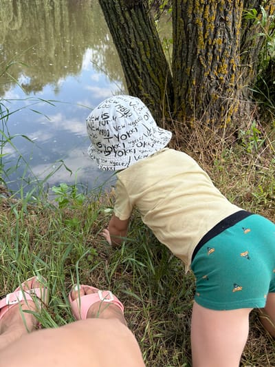 This is a vertical, close-up, eye-level shot of a child wearing a light yellow t-shirt, green shorts with a pizza pattern, and a white hat with black text, crouching on grassy ground next to a body of water. The child's hands are reaching towards the water. To the right of the frame, a person's legs and feet are visible, wearing pink sandals. A large tree trunk is on the left side of the image, with moss and lichen on its bark. The water in the background reflects the sky and trees, suggesting it is daytime with some clouds. The overall setting appears to be a natural, outdoor environment, likely near a river or lake. The text visible on the child's hat includes words like "LOS," "Ngeles," "New 1980," and "YOKKI."