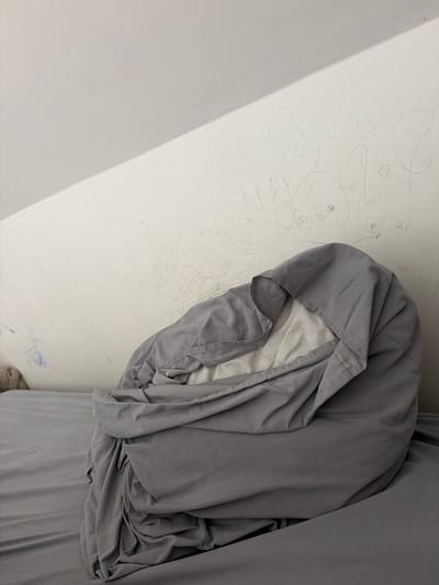 The image depicts a bed with rumpled grey bedding and a white mattress cover. The bedding is gathered into a large mound in the center of the frame. A white wall with child-like drawings in pencil and crayon serves as the background. The wall is textured and shows signs of wear. The overall scene suggests a domestic setting, likely a bedroom. The location is specified as Falenty Nowe, Poland.