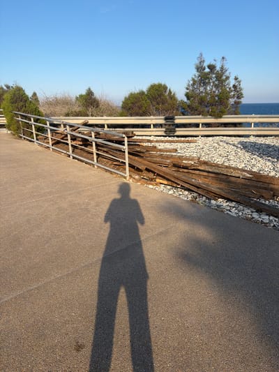 In Pervolia, Cyprus, a shadow of a person with legs and torso is cast on a textured, light brown paved surface. To the left and extending into the background, a metal railing with horizontal bars follows an upward slope. Beyond the railing, a pile of weathered wooden planks lies on a bed of light-colored gravel. In the background, green bushes and trees are visible, with a clear blue sky above. In the far distance, the blue sea meets the horizon.