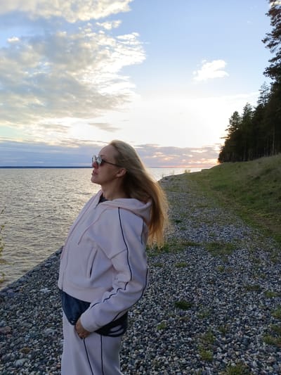 A woman stands on a gravel path alongside a body of water. She is wearing a light pink sweatsuit with black stripes and dark sunglasses. Her blonde hair is blowing in the wind. In the background, there's a forest on a grassy hill, the sky is bright with the setting sun and some clouds. The water is reflecting the sky's colors. Based on the information provided, the location is Вичелово, Russia.