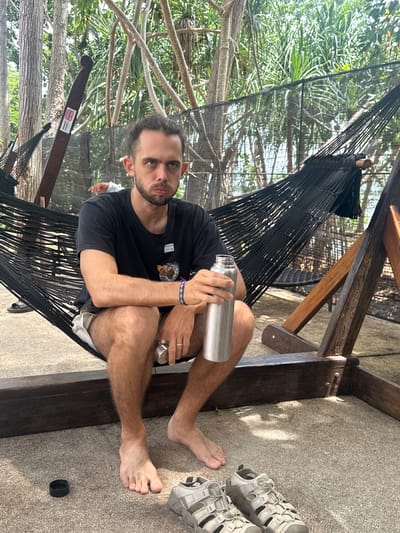 A man is sitting in a black hammock outdoors, holding a silver metal water bottle. He is wearing a black t-shirt and light-colored shorts. His feet are bare, and a pair of grey sandals are visible on the ground next to him. The hammock is suspended from a wooden structure, and behind the man, palm trees and foliage are visible, suggesting a tropical setting. The ground appears to be covered in a textured material, possibly concrete or gravel. The lighting suggests it is daytime, and the weather appears to be clear and sunny. The man is looking towards the camera with a neutral expression. A small white sticker with red text is visible on the wooden support beam of the hammock. The text on the sticker appears to be in Thai and English, and may indicate a weight limit or safety instruction, though it is difficult to read clearly. The overall scene depicts a man relaxing in a hammock in a tropical environment.
