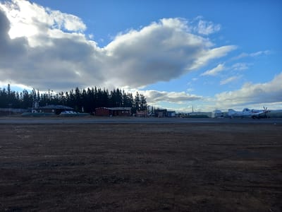 The image displays an airfield in Santa Juana, Chile, under a partly cloudy sky. The foreground consists of a broad expanse of dark brown, bare earth. In the midground, a line of dense, dark green coniferous trees runs horizontally across the left side. Situated in front of these trees are several structures: a light-colored building with a dark roof, a white vehicle parked nearby, and a reddish-brown building with a dark roof. A tall, dark mast or tower is visible behind the reddish-brown building. Extending from the center to the right midground is a paved runway or tarmac. On this surface, a white single-engine turboprop aircraft with red markings and a visible propeller is oriented toward the right. A dark ground support vehicle is positioned near the aircraft. Further in the background, several light-colored hangars or industrial buildings are adjacent to the tarmac. The sky above transitions from dense, grey clouds on the left to clearer blue with scattered white clouds on the right. No individuals are discernible within the scene.