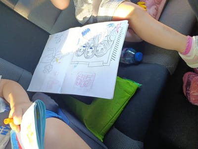 The image shows a close-up of a person's legs and an open coloring book inside what appears to be the back seat of a vehicle. The coloring book is on a green cushioned surface. The coloring book has drawings of people and some smudges of blue and red ink. A plastic bottle of water is also visible. The environment is dimly lit, suggesting it is inside a car. There are no visible landmarks or clues that indicate the location is in Svobodinovo, Bulgaria.