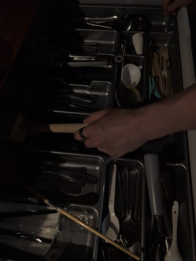 The image shows a close-up, overhead view of an open kitchen drawer filled with various utensils. A person's hand, wearing a ring on the right index finger, is visible reaching into the drawer. The drawer is organized with compartments for different types of cutlery and other kitchen tools. Items present include spoons, forks, knives, a small white funnel, a small white cup, and a wooden spatula or stirrer. The drawer appears to be made of dark gray plastic. The background is dark, suggesting the photo was taken in low light conditions. The scene is indoors, and based on the provided information, it is located in Słupsk, Poland. The overall impression is one of everyday domestic activity.