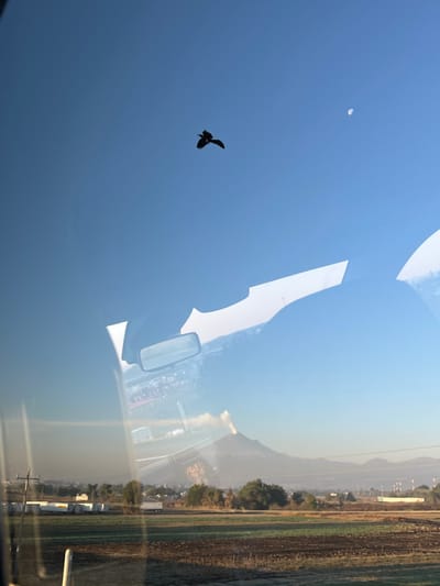 The image depicts an outdoor scene near Santa Ana Xalmimilulco, Puebla, Mexico, observed from inside a vehicle. The sky, predominantly clear and blue, features a dark, silhouetted bird in mid-flight with wings extended, located in the upper-center. A distinct crescent moon is visible in the upper-right quadrant. The horizon is dominated by the Popocatépetl volcano, actively emitting a visible plume of white smoke or ash from its summit. Below the volcano, the landscape includes distant hazy mountains, scattered vegetation, and faint structures indicative of a town or industrial area. The immediate foreground outside the vehicle shows an agricultural field with patches of green foliage and brown soil. White rectangular objects, possibly storage units or trailers, are situated on the left side of this field. The view is partially obscured by significant reflections on what appears to be a vehicle's windshield, including a central rearview mirror reflecting indistinct lights and structures, and white, irregular shapes consistent with the vehicle's interior elements such as the headliner or dashboard. A vertical white post, part of the vehicle's frame, is visible in the lower-left corner.