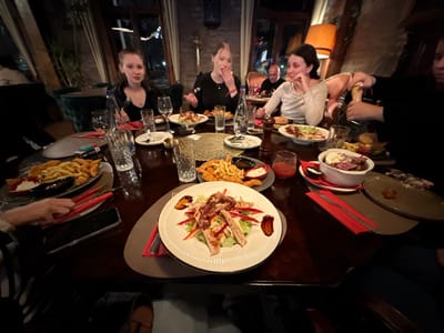 The scene depicts an indoor nighttime gathering at a restaurant or bar in Cuprija Municipality, Serbia. Approximately seven to eight individuals are seated around a large, dark wooden table, engaged in dining and social interaction under warm, dim ambient lighting.
In the left mid-ground, a young female with long, dark hair wears a black top and looks directly forward. To her right, a female with blonde hair, dressed in a dark top, has her hand near her mouth, potentially speaking or pausing. A red Marlboro cigarette packet is positioned on the table adjacent to her. Further right, another female with dark hair, wearing a light-colored long-sleeved top, gestures towards the right. In the background, a male with dark hair is visible seated. On the far right, a partially visible individual is operating a dark wooden salt or pepper grinder over a dish. A person's arm with a striped sleeve and a smartphone are visible in the left foreground.
The table is laden with multiple dishes: a prominent oval plate in the foreground holds a grilled chicken salad comprising chicken strips, lettuce, red bell peppers, and bacon. Other plates contain fried chicken tenders or nuggets accompanied by french fries and small bowls of dipping sauces. A white bowl holds sliced meat, possibly pastrami or roast beef, with a creamy topping. Beverages include several clear glasses containing water or other drinks, a small glass with a red liquid, and a brown glass bottle. Red fabric napkins, silverware, and a large dark wooden pepper mill are also on the table.
The environment features exposed brick walls, dark-framed windows reflecting the night, and dark green velvet armchairs in the background. A tall potted plant stands on the left. An orange-shaded lamp is visible in the right background, contributing to the subdued lighting. Wooden architectural elements are present overhead.