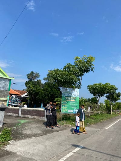 The scene is a street in Kecamatan Tulangan, Indonesia, located at coordinates -7.474 latitude and 112.621 longitude.  Four men, all wearing long-sleeved shirts and pants, are walking on the street. One man is wearing a black headscarf and white long-sleeved shirt. Another is wearing a white headscarf and black long-sleeved shirt. The other two men are wearing white head coverings. A small boy with short hair, wearing a blue shirt and shorts, is also walking on the street. The men are walking on a paved street with a white line down the center. To the left of the street is a low brick wall. Behind the wall is a green house with a green roof. There is a large, green sign to the right of the men, advertising "JUM'AT BARU" (New Friday) with information about religious events. Behind the sign are large trees with green leaves. The sky is blue and mostly cloudless.  The men are walking in the same direction on the paved road.