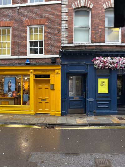 An exterior forensic view depicts two adjacent shopfronts and a section of a street in York, United Kingdom, specifically at 55 Stonegate. The scene is static, devoid of people or overt activity.
The building on the left features a ground floor entirely painted in a bright yellow hue. It houses a L'Occitane en Provence store, indicated by "L'OCCITANE EN PROVENCE" signage above the display window and the central six-panelled wooden door. The door is marked with the address "55" above it and includes a brass letterbox. The large display window to the left of the door showcases various products and a large poster featuring a person. Above the yellow ground floor, the first story is constructed of reddish-brown brickwork and features two white-framed sash windows, behind which yellow interior blinds or coverings with "L'OCCITANE EN PROVENCE" text are visible. A small blue triangular alarm box is mounted on the brickwork between floors.
The building on the right presents a deep navy blue ground floor facade. This section includes a recessed blue wooden door, marked with "53" above it, leading into a premises. To its right, a window area displays a bright yellow rectangular sign proclaiming "THE SET MENU." Directly above this blue shopfront, a vibrant flower box brimming with pink, white, and purple blossoms is suspended. The first story of this building is also reddish-brown brick, featuring two white-framed sash windows with arched brick lintels, contrasting with a vertical lighter-colored brick or stone strip running up the corner where it meets an unseen adjacent building.
The street level consists of a grey flagstone pavement directly in front of the shops, which transitions to a wet, dark asphalt road. The road surface reflects ambient light, suggesting recent precipitation or damp conditions. Double yellow lines are visible along the curb, and a rectangular metal drain cover is situated in the gutter. A square metal utility cover is partially visible on the road further to the right.