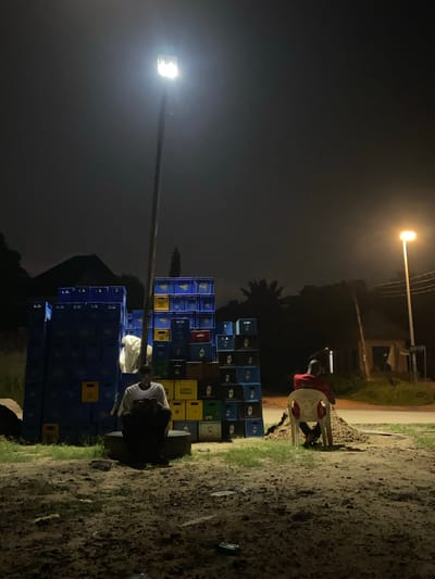 The image depicts a nighttime scene in Nguzu Edda, Nigeria. A large stack of blue, green, yellow, and black beer crates is centrally located. To the right, a person sits on a white plastic chair wearing a red shirt. Another person is squatting near the crates, dressed in a white t-shirt. Two street lights illuminate the area. The ground is dirt with patches of grass.