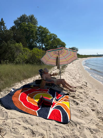 A man sits in a beach chair under a striped umbrella on a sandy beach in Hampton Bays, New York. He is wearing a light-colored baseball cap and light-colored shorts. In front of the chair, a colorful blanket with a circular pattern is spread on the sand. A black bag and a paper grocery bag are on the sand behind the chair. A beach towel hangs from the umbrella. The ocean is to the right of the man, with gentle waves lapping the shore. Behind the beach, there is a line of tall green grass and trees. The sky is clear and blue. A bird is visible flying in the distance.