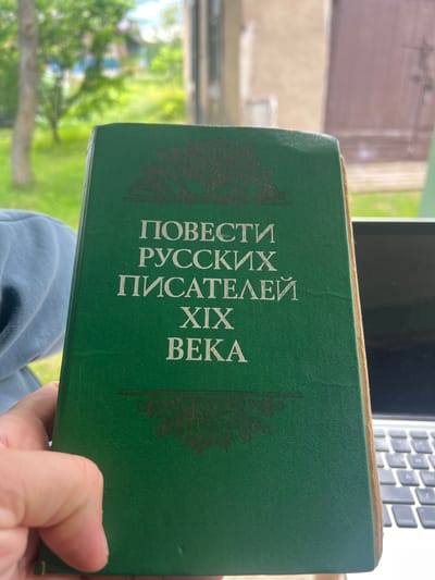 A green book with gold lettering in Russian is held by a person's left hand. The lettering on the cover reads "ПОВЕСТИ РУССКИХ ПИСАТЕЛЕЙ XIX ВЕКА," which translates to "Stories of Russian Writers of the 19th Century." The book is positioned in the foreground, obscuring the lower portion of a laptop keyboard. The background is blurred, showing a bright, grassy outdoor environment with trees, suggesting a sunny day. The image appears to be taken from a slightly elevated angle, looking down at the book. The location of Бучацінскі сельскі Савет, Belarus is not discernible from the visual information provided in the image.