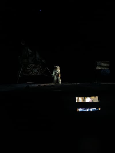 An astronaut stands on the moon's surface near the lunar lander. The American flag waves in the distance. Above the scene, a small blue point could represent Earth in the black sky. In the foreground, a screen displays a man's face and subtitles. The overall scene evokes the historical lunar landing, possibly a museum display.