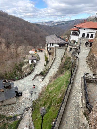 This aerial photograph captures a descending stone-paved pathway bordered by buildings, greenery, and lampposts, likely in the city of Kriva Palanka, North Macedonia. The pathway is flanked by black metal railings and occasional benches. A person dressed in black with red pants walks along the path. Buildings with white walls and red-tiled roofs line the path. Further into the background, trees and a mountain ridge can be seen. The sky features a combination of blue patches and heavy cloud cover. Stone structures, including what appears to be a well, are present at the bottom of the image.