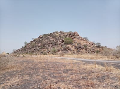 The image depicts a rocky hill in a dry, arid landscape, likely located near the city of Shahid Afandi in Sudan. The hill is primarily composed of large, rounded rocks and boulders, sparsely covered with dry vegetation and a few scattered trees. A paved road passes along the base of the hill. The foreground shows a field covered in dry grass and patches of dark soil, possibly from burning. The sky is clear and pale blue. There are no people visible in the image. A simple structure, possibly a hut or shelter, can be seen in the far left side of the image.