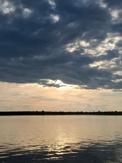 A body of water reflects the light of the setting sun, which is partially obscured by dark, dramatic clouds. The horizon is a dark silhouette of land with sparse trees. The scene appears to be a natural landscape, possibly a river or lake, near the village of Введеновка, Russia. There are no people or specific objects visible that would indicate human activity or urban environment. The water is calm with gentle ripples, and the sky is filled with a mix of dark and light clouds, creating a contrast between shadow and light.