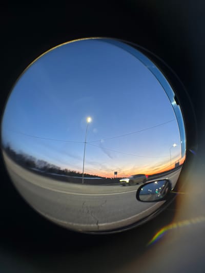 The image depicts a fisheye lens view of an evening scene. A blue sky with a street light dominates the left side of the frame, while a road with a car's side mirror is visible on the right. The horizon shows hues of orange and white, suggesting a sunset or sunrise. A dark line of trees or foliage runs along the bottom of the road. The location in Kuregovo, Russia, cannot be confirmed from the image.