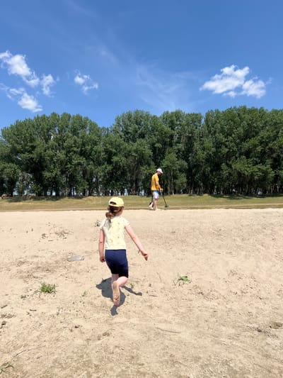 A young girl with a yellow hat and shirt, wearing dark blue shorts, walks barefoot across a sandy beach. Behind her, a man in a yellow shirt and white hat uses a metal detector. A line of tall green trees forms a backdrop against a bright blue sky with scattered white clouds. The location is the sandy riverbank in Indiya, Moldova.
